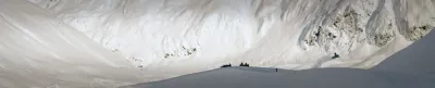 A wide panoramic view of a massive snow-filled bowl in Alaska with snowmobilers in the distance.
